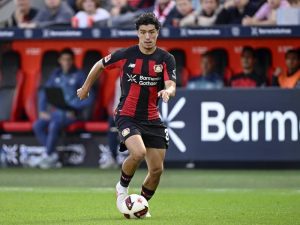 Bayer Leverkusen player in black and red striped kit dribbling the ball on the pitch during a Bundesliga match, with Barmenia Gothaer advertising behind him.