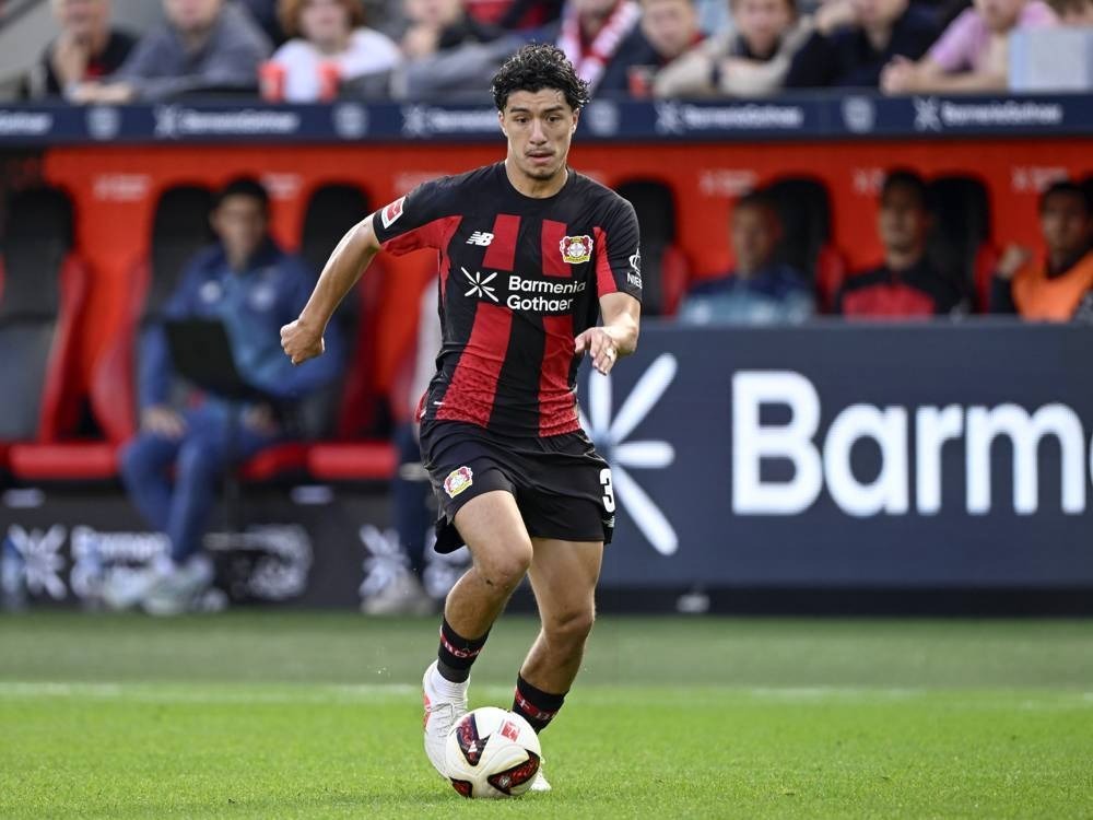 Bayer Leverkusen player in black and red striped kit dribbling the ball on the pitch during a Bundesliga match, with Barmenia Gothaer advertising behind him.