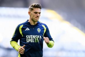 A football player in a navy and yellow Svenska Spel training top with the Swedish crest, jogging in a stadium.