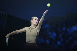 Tennis player in a khaki sleeveless top and brown skirt serving a yellow ball under stadium lights, with a crowd blurred behind her