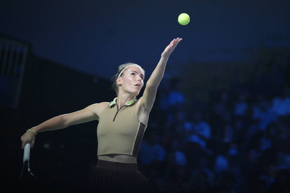 Tennis player in a khaki sleeveless top and brown skirt serving a yellow ball under stadium lights, with a crowd blurred behind her