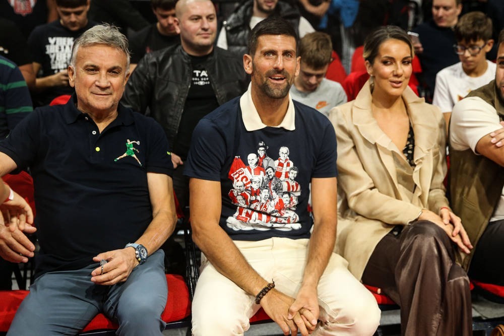 Man in dark navy T-shirt with a red-and-white graphic, seated courtside at a sporting event beside an older man in a polo and a woman in a beige coat