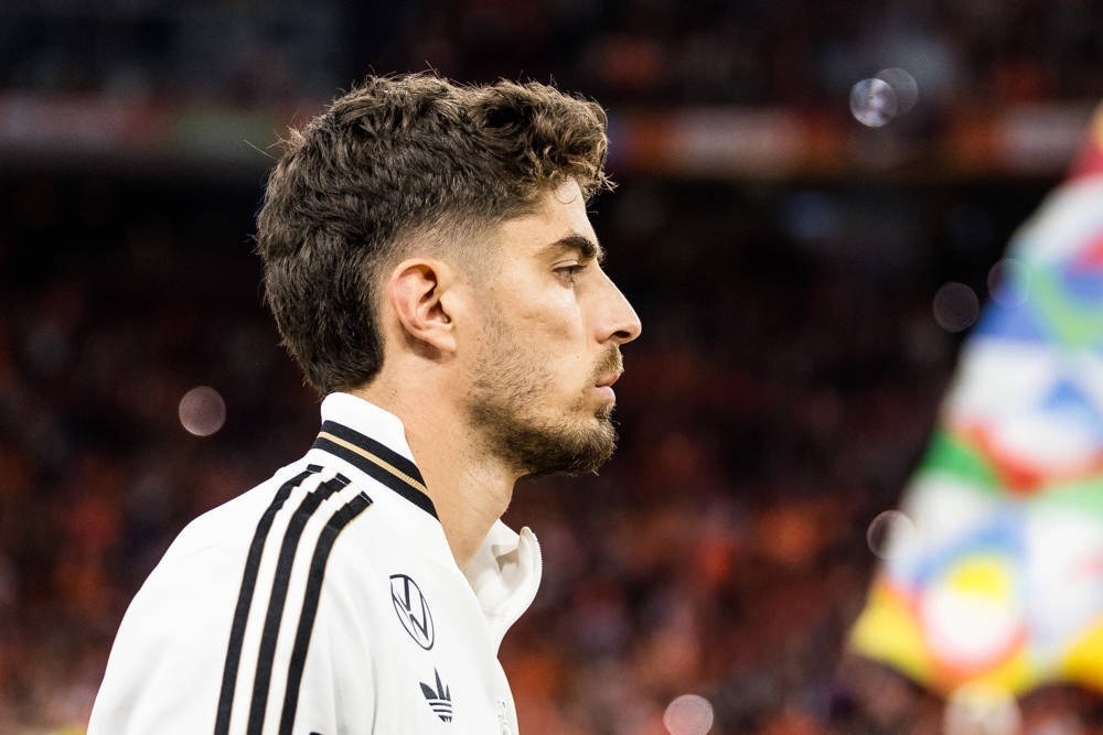 A man with curly dark hair and a short beard in a white Adidas jacket with black stripes and a Germany crest, in profile at a stadium.
