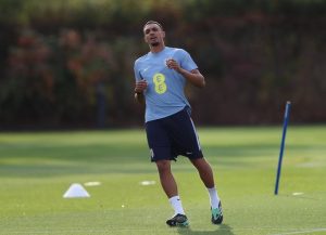A football player in a light blue training shirt with a yellow EE sponsor logo and navy shorts, jogging on a green training pitch past a blue marker pole.