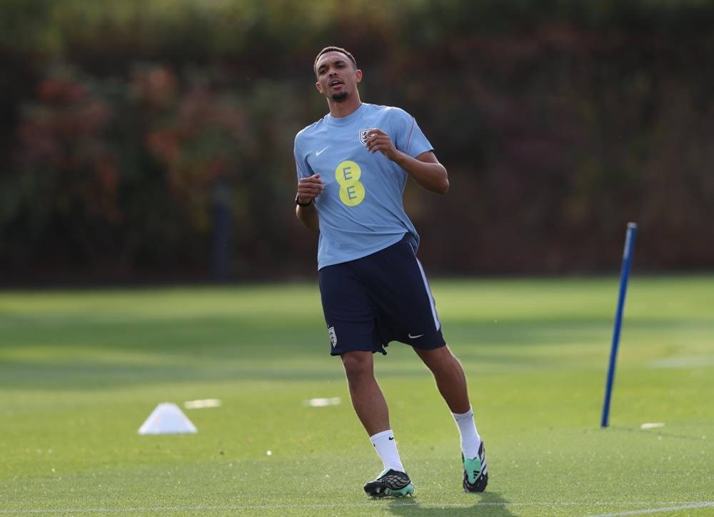 A football player in a light blue training shirt with a yellow EE sponsor logo and navy shorts, jogging on a green training pitch past a blue marker pole.