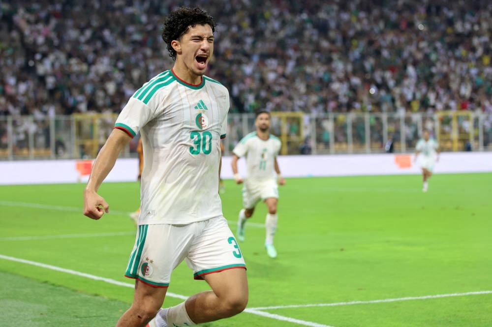 Young footballer in white and green Algeria kit, number 30, shouting and clenching fist in celebration on a stadium pitch.