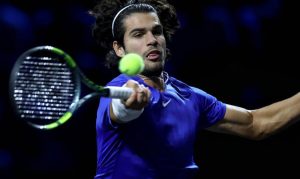 Tennis player in a blue Nike shirt and white headband hits a forehand return with a green-and-black racket at a night event.