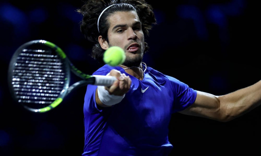 Tennis player in a blue Nike shirt and white headband hits a forehand return with a green-and-black racket at a night event.