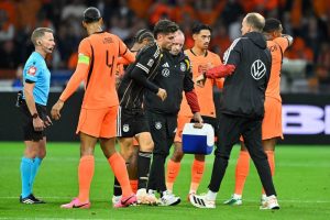 Germany player receiving treatment and helped off the pitch by staff in black kit, as Netherlands players in orange look on, Nations League match
