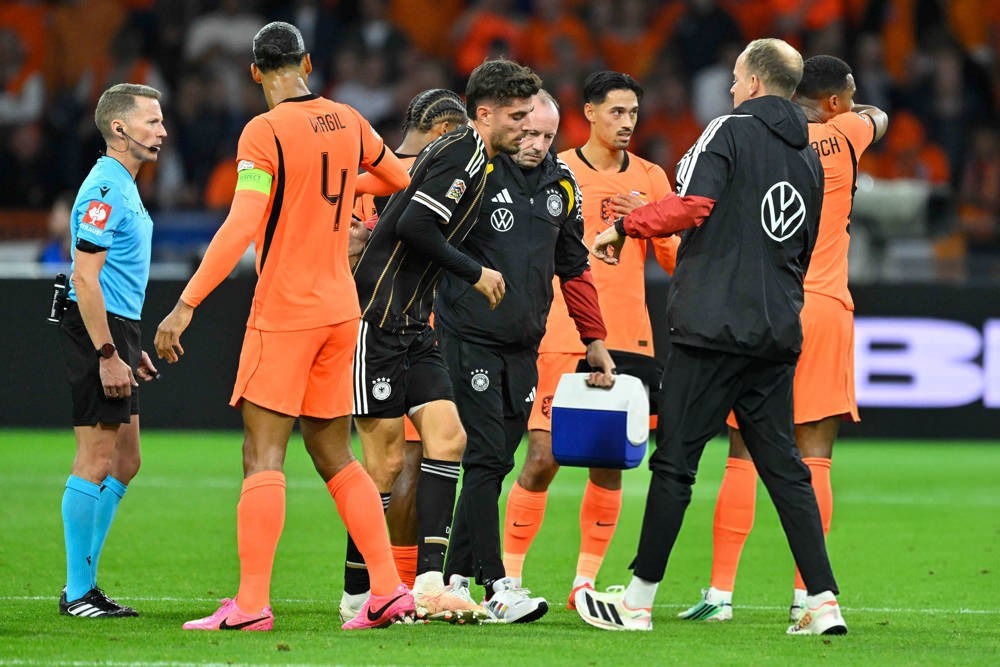 Germany player receiving treatment and helped off the pitch by staff in black kit, as Netherlands players in orange look on, Nations League match