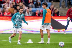Bernardo Silva in teal Portugal training kit warms up on a pitch, with teammate in orange bib nearby and a ball on the grass.