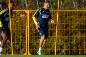 Football player in navy Svenska Spel training kit running on a pitch beside yellow fencing, with another player in the background. Sweden national team train…