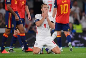 England footballer wearing number 9 and captain's armband kneels on the pitch, hands pressed to his face, while Spain players in red stand behind him.