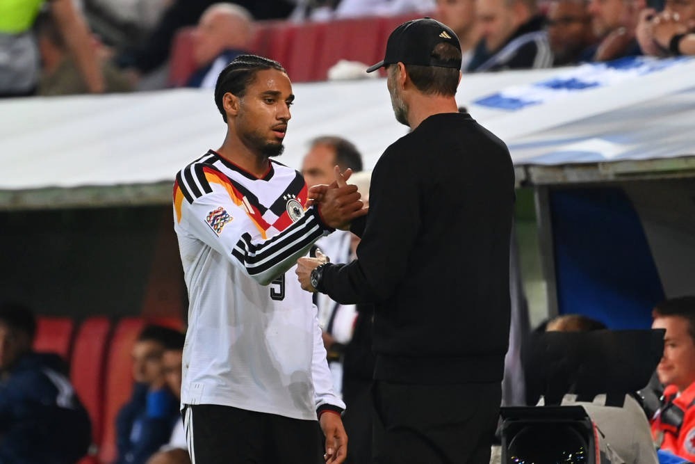 Football player in white and black Germany kit with braided hair shaking hands with a coach in a black cap and jacket on the touchline during a match.