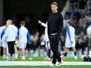 Man with grey beard in black sweatshirt and trousers, hands in pockets, standing on a pitch before a match, players warming up behind him.