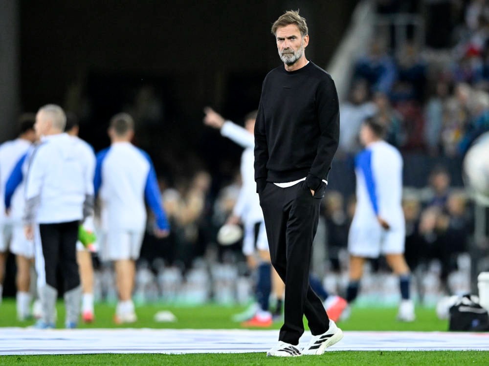 Man with grey beard in black sweatshirt and trousers, hands in pockets, standing on a pitch before a match, players warming up behind him.