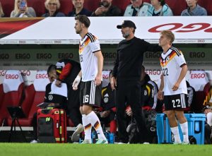 Two German players in white home kits with black shorts walk past a bench as a man in a black cap and tracksuit looks on, during a match at a stadium.