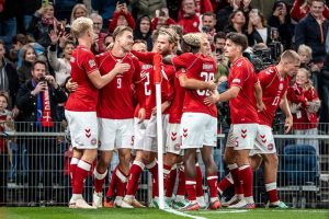 Denmark players in red kits celebrate a goal in front of fans, number 9 and teammates embracing near the corner flag at a Nations League match