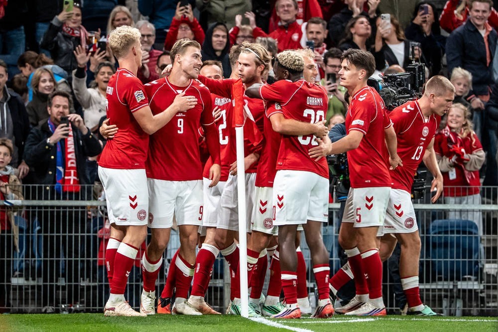Denmark players in red kits celebrate a goal in front of fans, number 9 and teammates embracing near the corner flag at a Nations League match