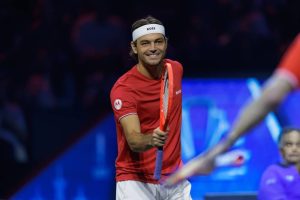 Taylor Fritz smiling in a red Team World shirt and white headband, holding an orange racket at the Laver Cup, with a blurred teammate in the foreground.
