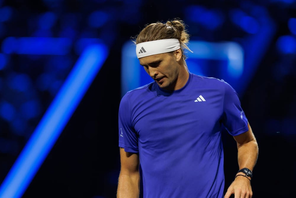 Alexander Zverev in blue Adidas shirt and white headband, head bowed on court under blue arena lights at the Laver Cup