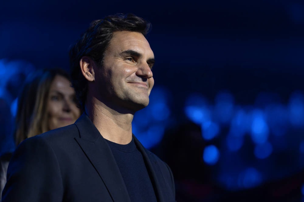 Man in dark blazer and black shirt smiling on a blue-lit stage, with a woman with blonde hair behind him