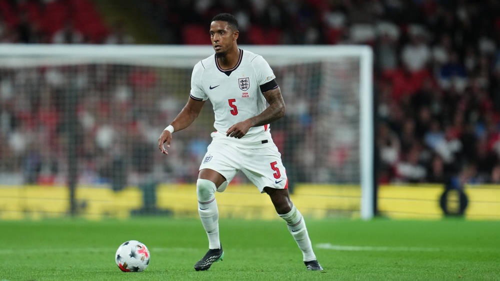 Football player in white England kit, number 5, dribbling the ball on a green pitch at a stadium during a match.