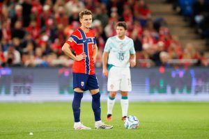 Football player in red and navy Norway kit with hands on hips on a pitch, another player in white Portugal kit behind him with ball, stadium crowd behind