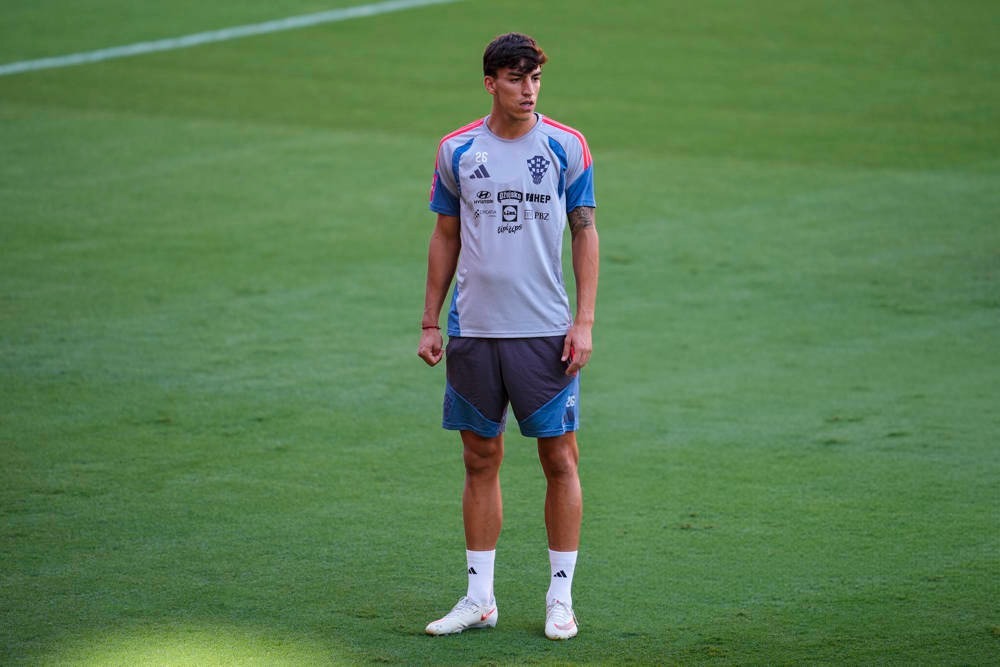 Young male footballer in grey training kit and grey shorts standing on a green pitch, looking ahead during a training session