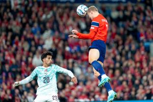 Player in orange Norway kit with number 9 heads the ball during a match, opponent in white Portugal kit number 20 nearby
