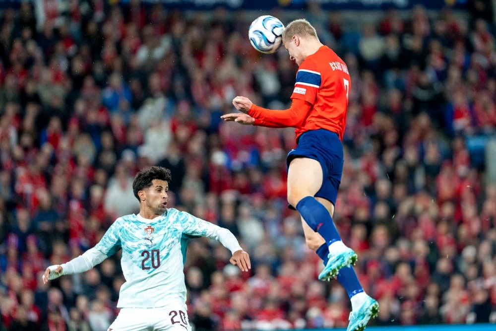 Player in orange Norway kit with number 9 heads the ball during a match, opponent in white Portugal kit number 20 nearby