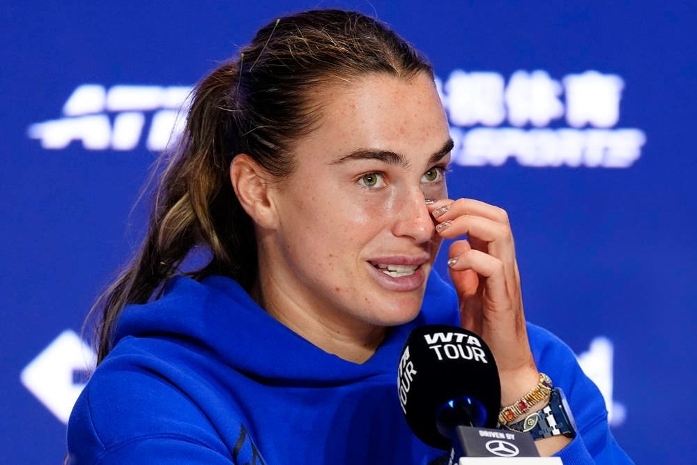 Tennis player in a blue hoodie wiping her eye at a WTA Tour press conference, with a Mercedes-Benz mic flag in front of her.