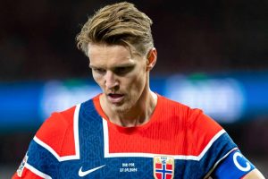 Footballer in red and blue Norway kit with captain's armband and crest, looking down during the NOR-POR Nations League match.