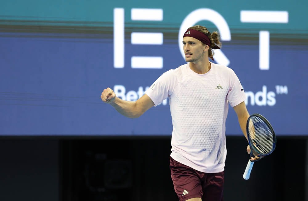 Alexander Zverev celebrates with a fist pump on court during a match, holding his racquet, with a sponsor backdrop behind him.