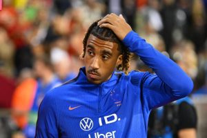 Football player in blue training top with Volkswagen ID and Uber logos, hand on head, looking aside at a stadium crowd. Article likely about Leny Yoro.