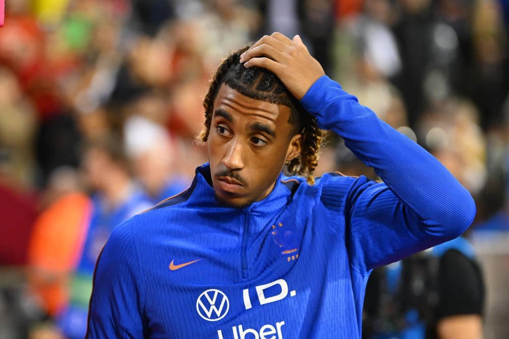 Football player in blue training top with Volkswagen ID and Uber logos, hand on head, looking aside at a stadium crowd. Article likely about Leny Yoro.