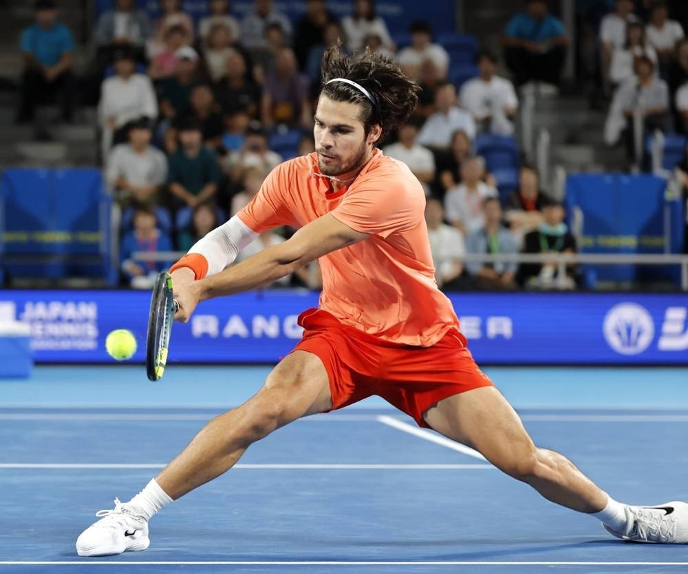 Tennis player in orange shirt and red shorts lunging to hit a forehand on a blue court at the Japan Open, with spectators behind him.