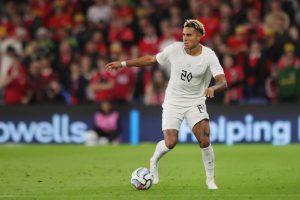 Footballer in a white kit with number 20 and yellow-blond hair dribbling a ball on a green pitch, with red-clad fans behind. Likely Antonio Nusa.