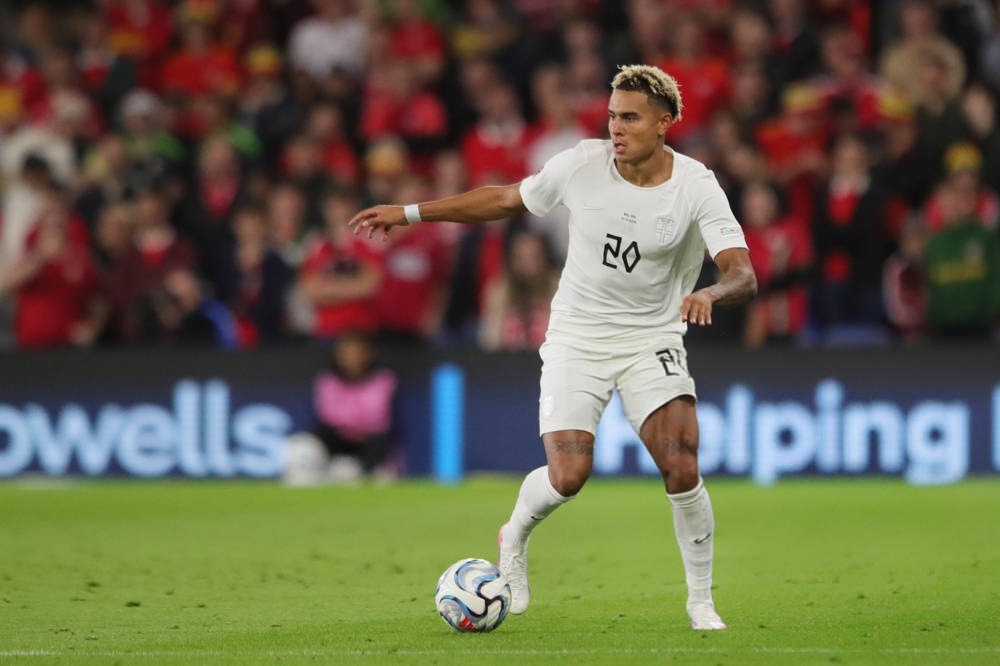 Footballer in a white kit with number 20 and yellow-blond hair dribbling a ball on a green pitch, with red-clad fans behind. Likely Antonio Nusa.