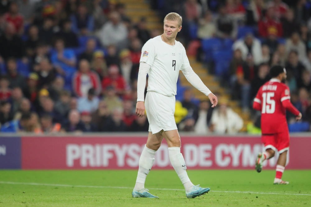 Football player in a white kit with number 9 walking on a pitch, a Wales player in red behind him at a Cymru stadium.