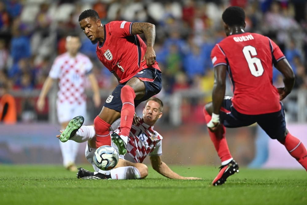 England player in red kit battles for the ball with a Croatia player in red-and-white checks during a match. Teammate "Guehi" number 6 watches on.