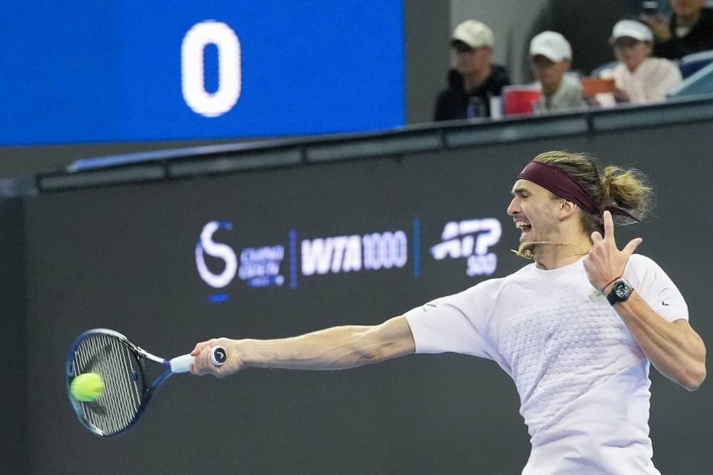 Alexander Zverev hits a forehand return at a tournament, wearing a white shirt and maroon headband, with a yellow ball in play.