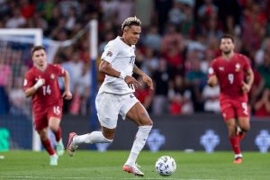 Antonio Nusa in a white kit dribbling a ball past red-shirted Serbian players during a match in front of a crowd.