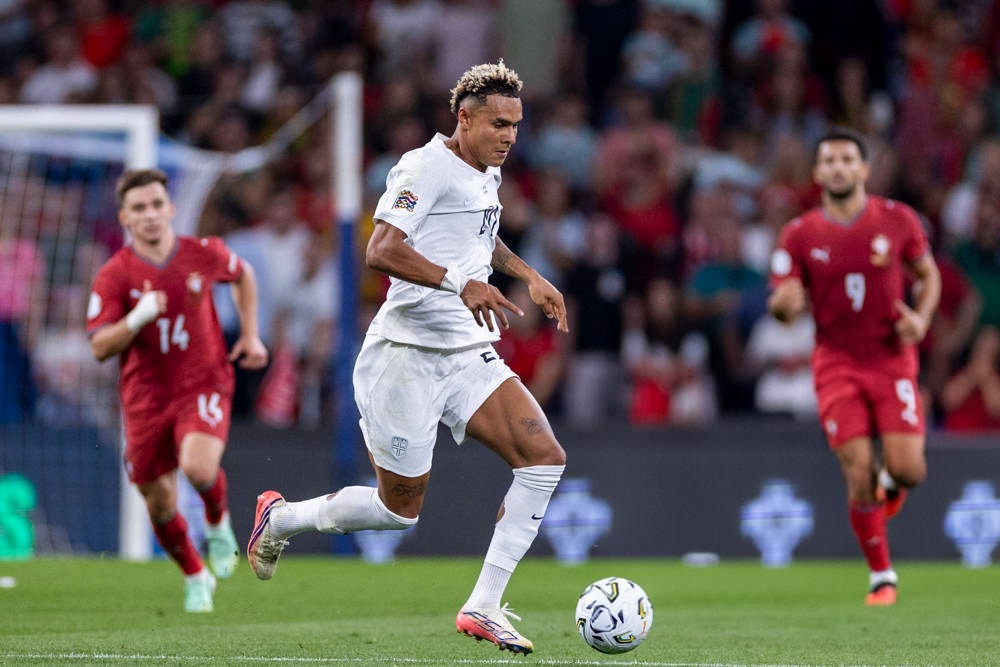 Antonio Nusa in a white kit dribbling a ball past red-shirted Serbian players during a match in front of a crowd.
