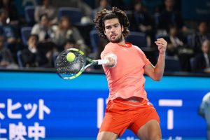 Tennis player in orange shirt and shorts hitting a forehand with a Babolat racket at an indoor event, Japanese signage behind him.