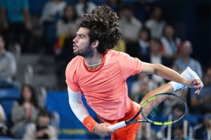Carlos Alcaraz in a coral shirt mid-serve at a tournament, racket extended, crowd blurred behind him