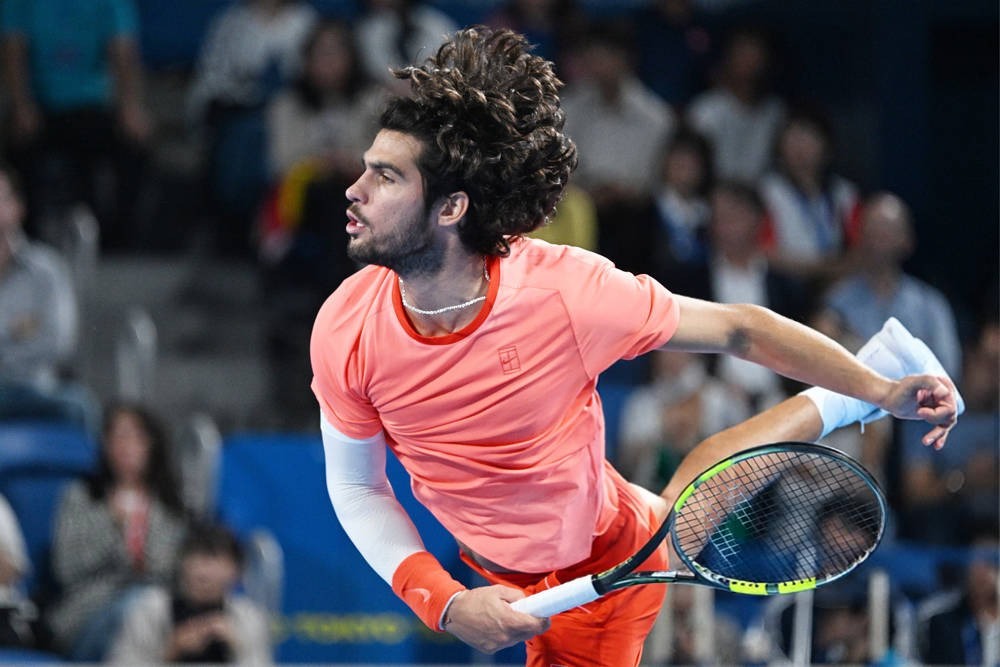 Carlos Alcaraz in a coral shirt mid-serve at a tournament, racket extended, crowd blurred behind him