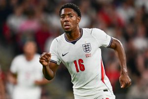 Footballer in white England No. 16 kit with three lions crest, running during a match in a blurred stadium crowd