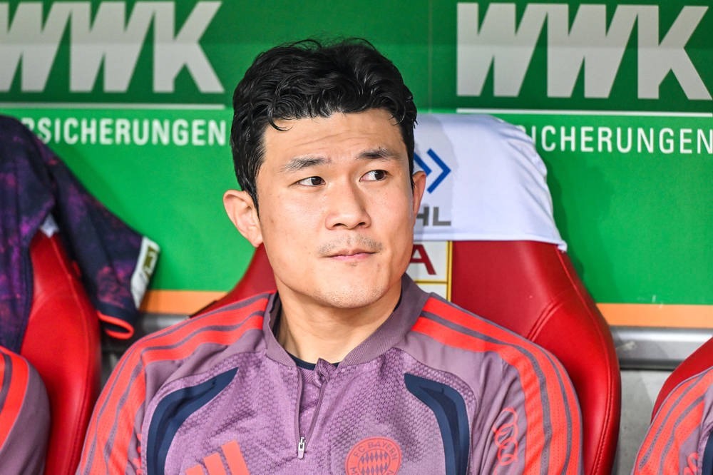 Man seated on a red bench in a purple Bayern Munich training top, looking to the side in the team dugout.