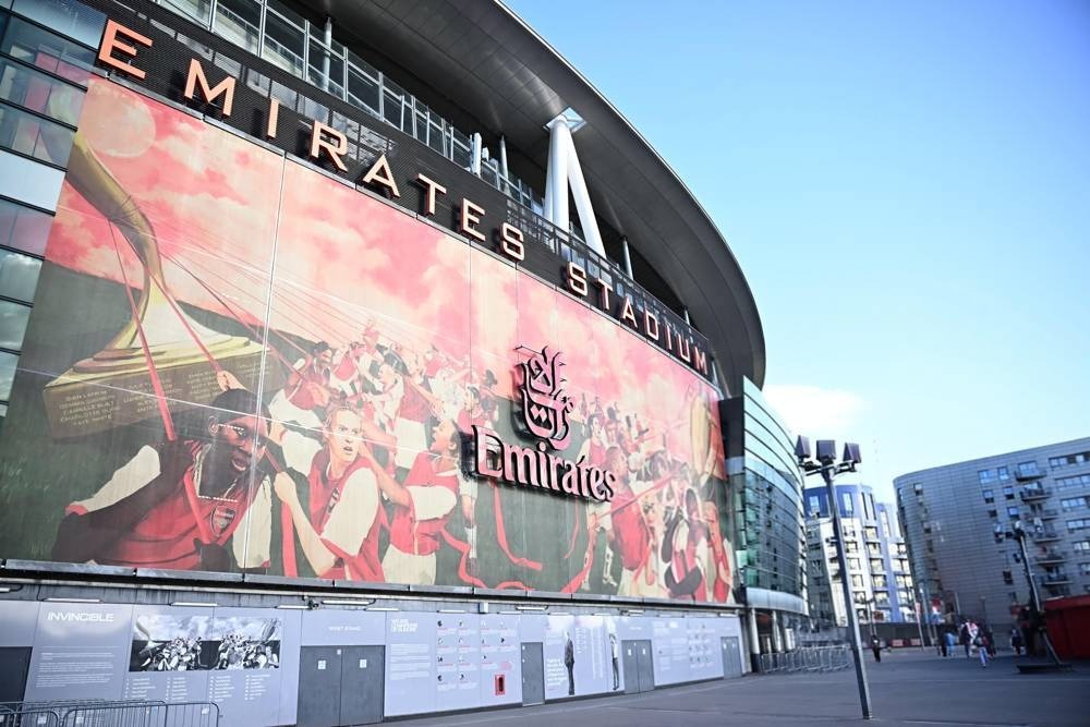 Emirates Stadium exterior in London, with a large red-and-white mural of Arsenal players and the Emirates sponsor logo on its facade, under a clear blue sky.
